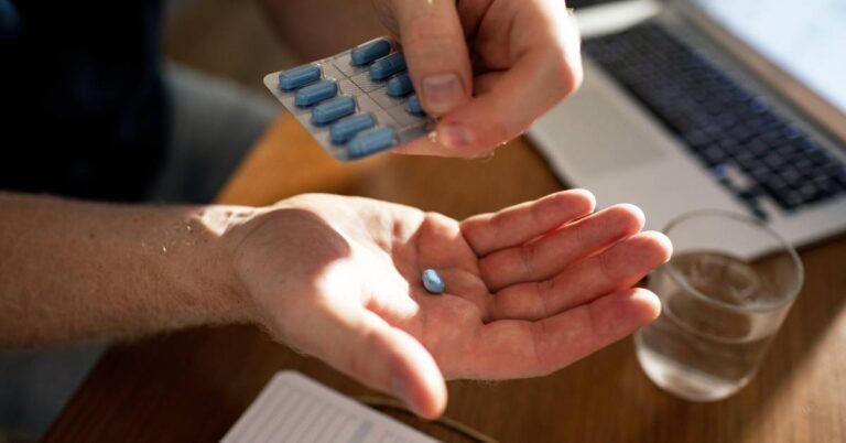 A man's hand holds a blue pill over a desk that has a glass of water, a laptop, and a desk calendar sitting on it.