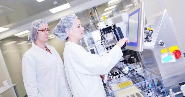 Two women working at a lab stand at a machine while one of them presses buttons on its screen. A glass wall is behind them.