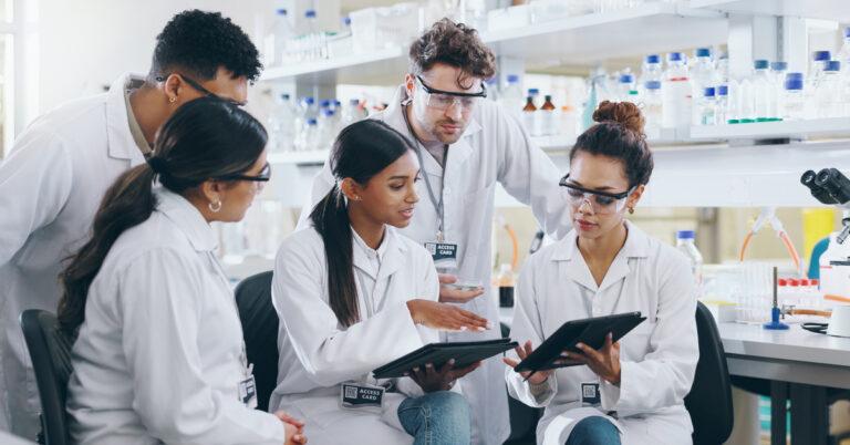 A group of scientists is discussing issues together in a lab. Some of the scientists are sitting, and others are standing.