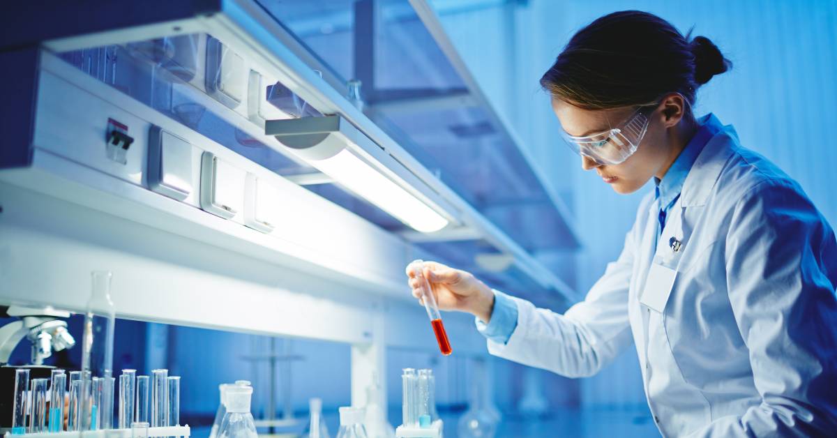A lab technician looks at a red chemical in a tube. She wears a white lab coat and safety glasses as she works.