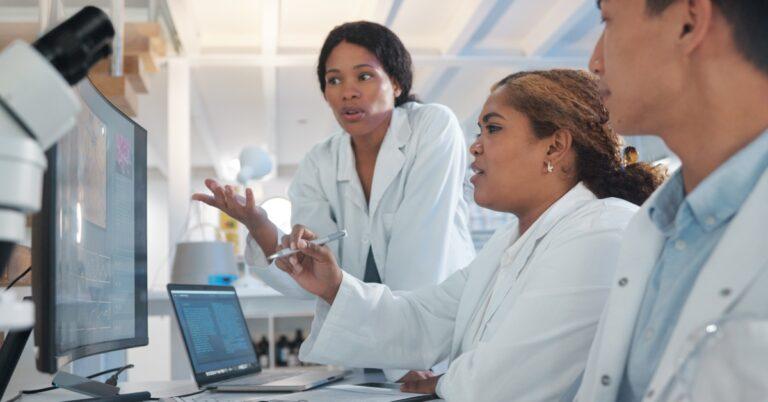 Three lab employees sit together at a counter and look at the information on a computer screen. They also have a laptop.