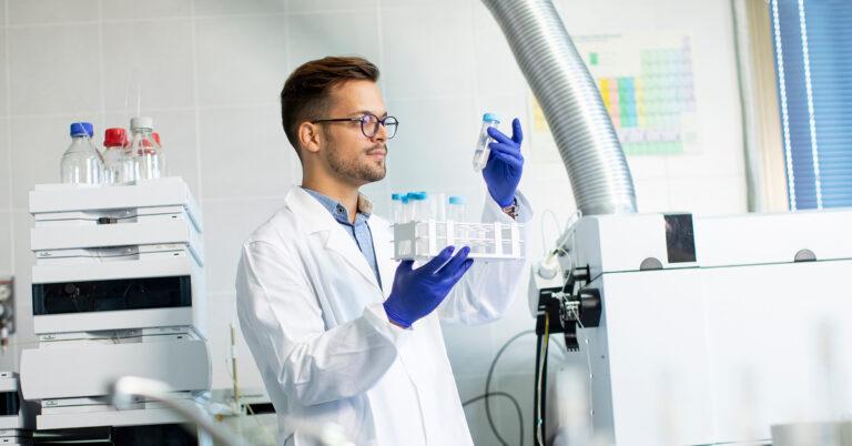 A lab employee holds a container of chemical samples in one hand and a test tube of one in the other.