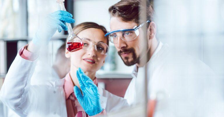 Two scientists stand in a lab, looking at a red chemical in a long-necked glass container. Both wear safety glasses.