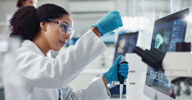 A woman working in a lab holds a glass vial in one hand an a dropper in the other. She wears safety glasses.