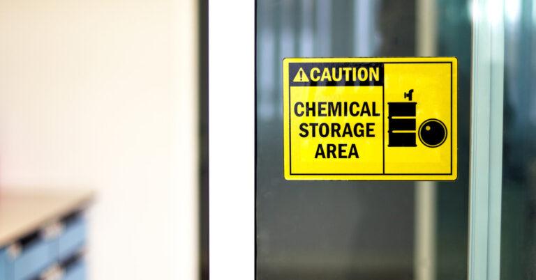 A yellow sign on a glass door in a workplace reads "Caution -- Chemical Storage Area." It features two black barrels.