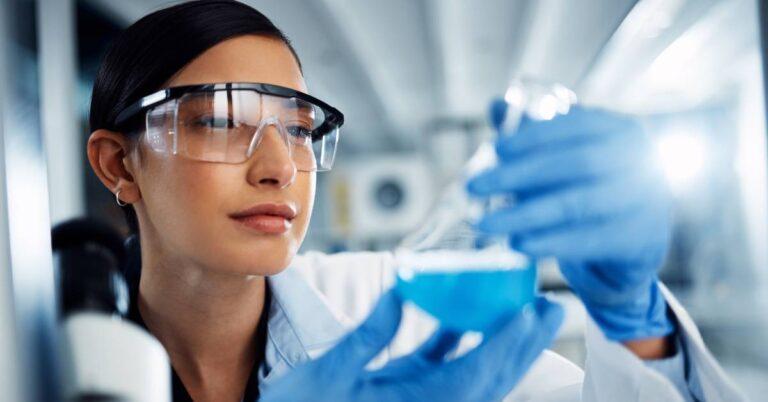 A lab scientist smiles as she looks at a blue chemical liquid in a glass flask. She wears blue plastic gloves.