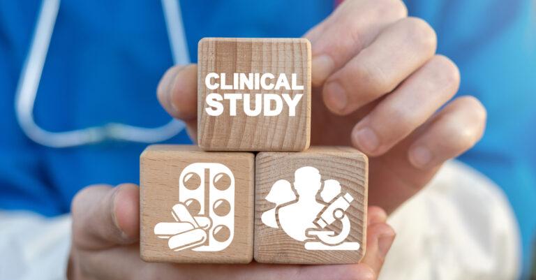 A researcher holds three wooden blocks in their hand, including one that reads "Clinical Study." They wear a blue shirt.