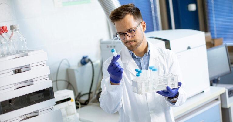 A researcher closely looks at a sample in his hand while holding more in his other hand. He is wearing glasses.